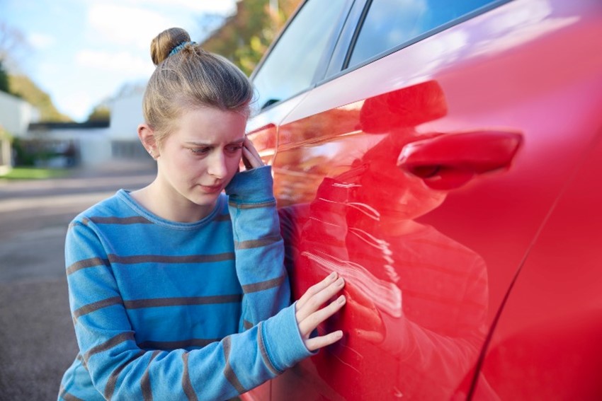 Young worried teenage female driver looking at damaged car after accident